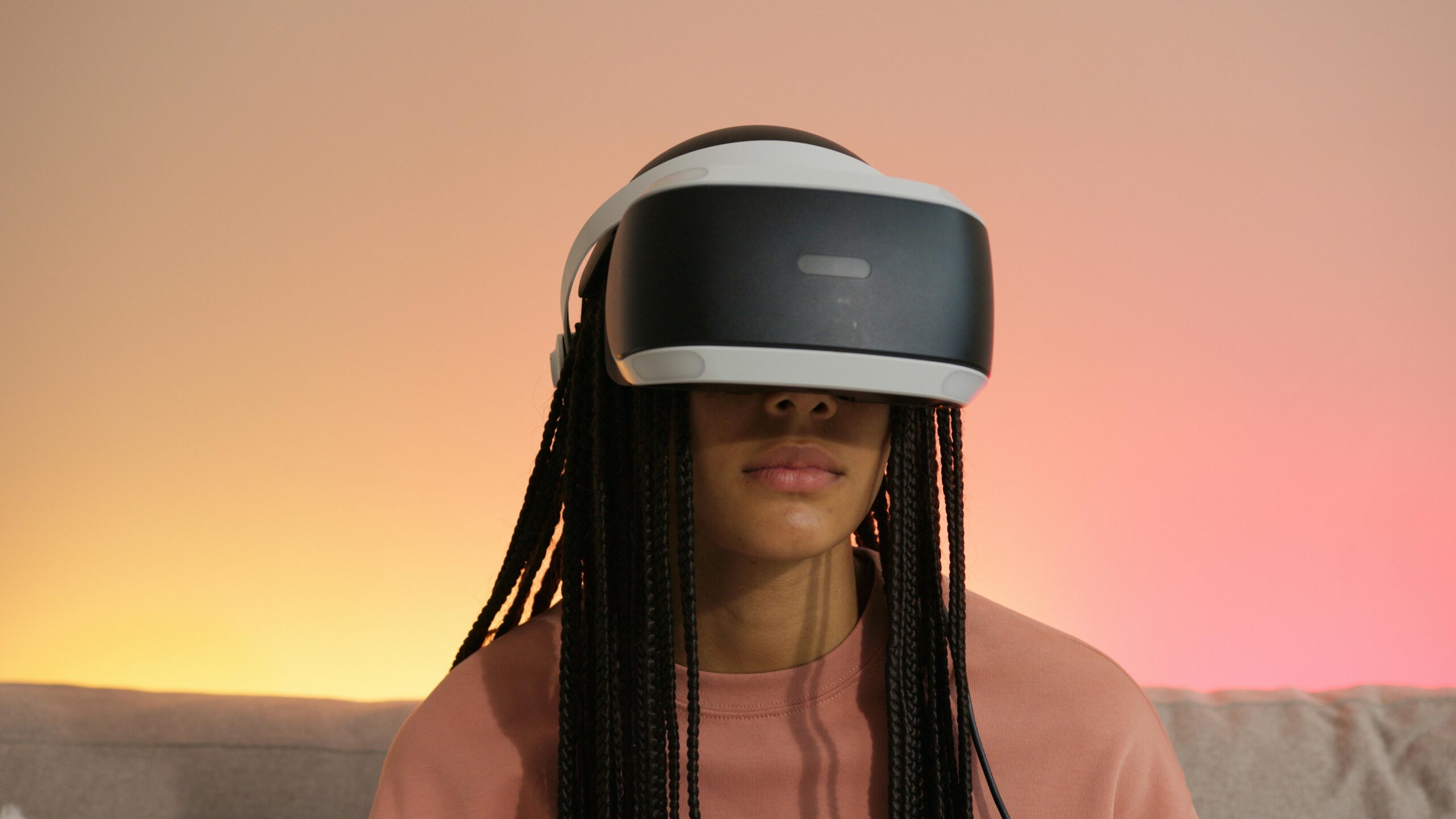 Woman with braided hair wearing a modern VR headset, set against a soft pink sunset background, illustrating the immersive experience of virtual reality.