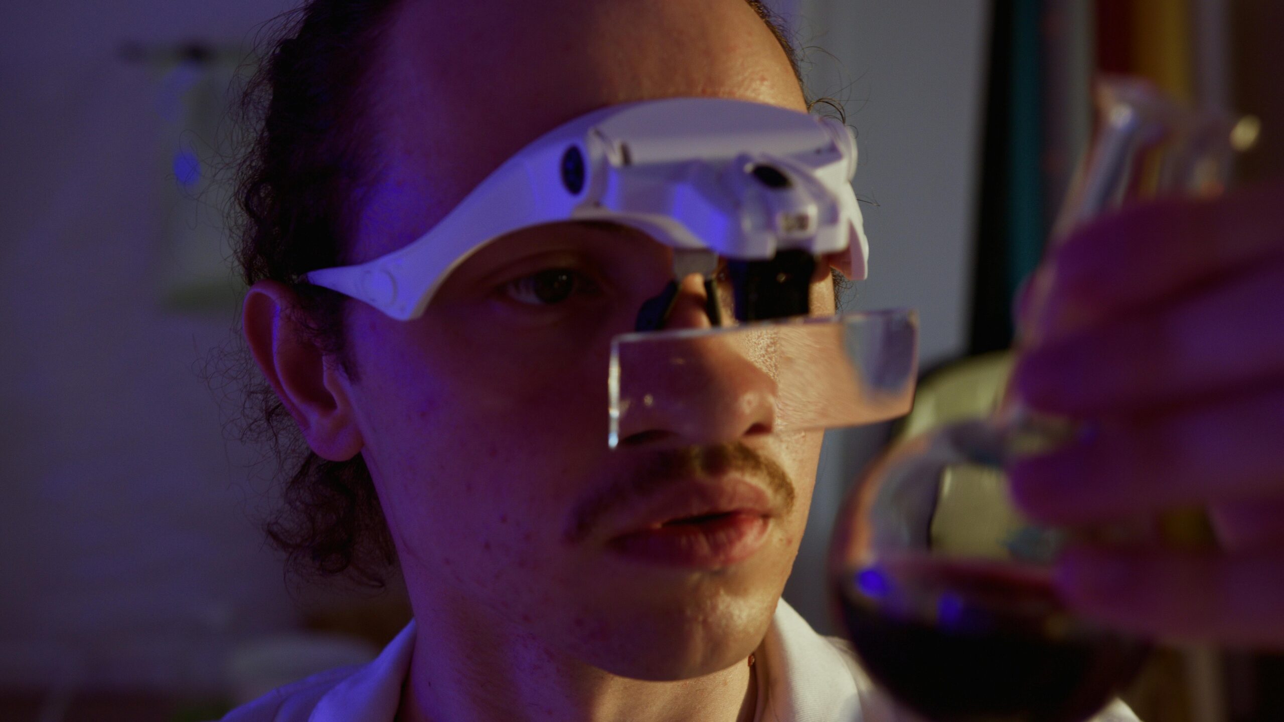 A close-up of a scientist wearing a futuristic head-mounted display and examining a glass of dark liquid, reflecting the integration of advanced technology in biotechnology.
