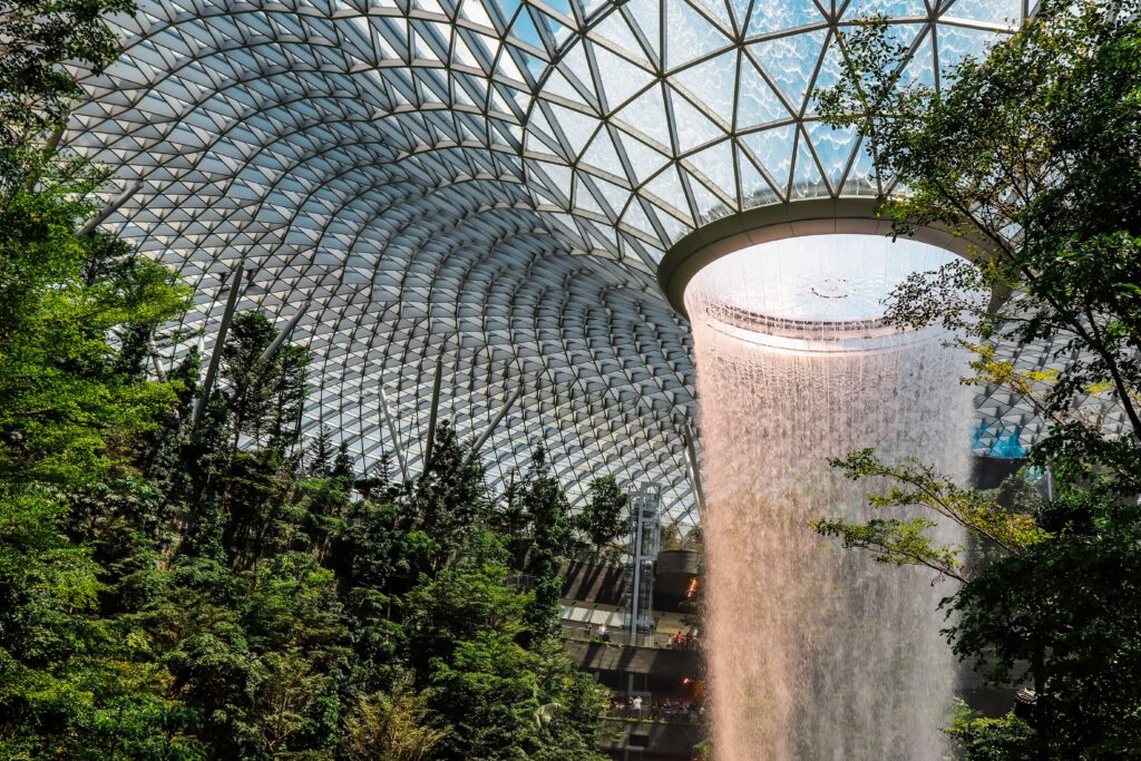 Indoor waterfall green tech cascading down a circular structure in the Jewel Changi Airport, Singapore, surrounded by a lush green forest and a futuristic glass and steel dome.