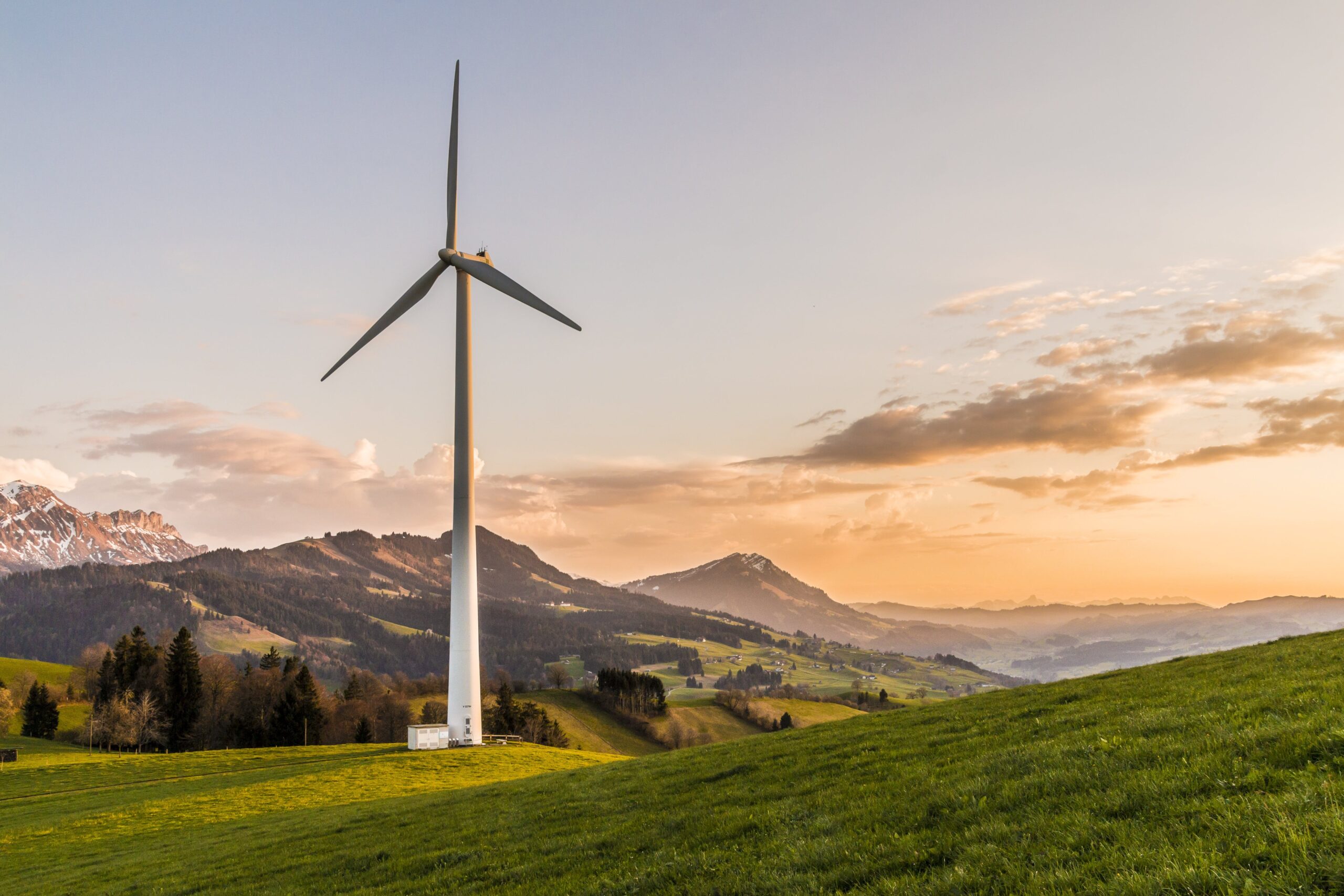 Solitary wind turbine on a green hill during sunset with the Swiss Alps in the background, symbolizing sustainable energy and environmental conservation.